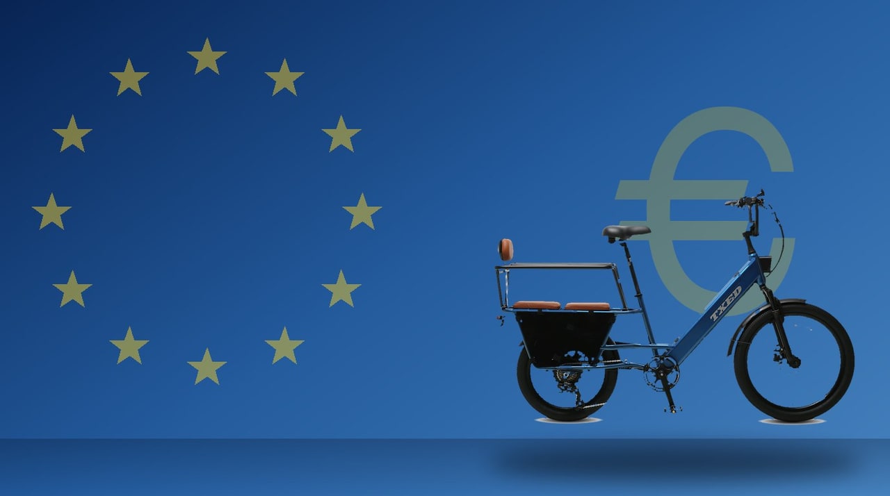Blue TXED cargo e-bike in front of a European Union flag inspired blue background with gold stars and a euro symbol